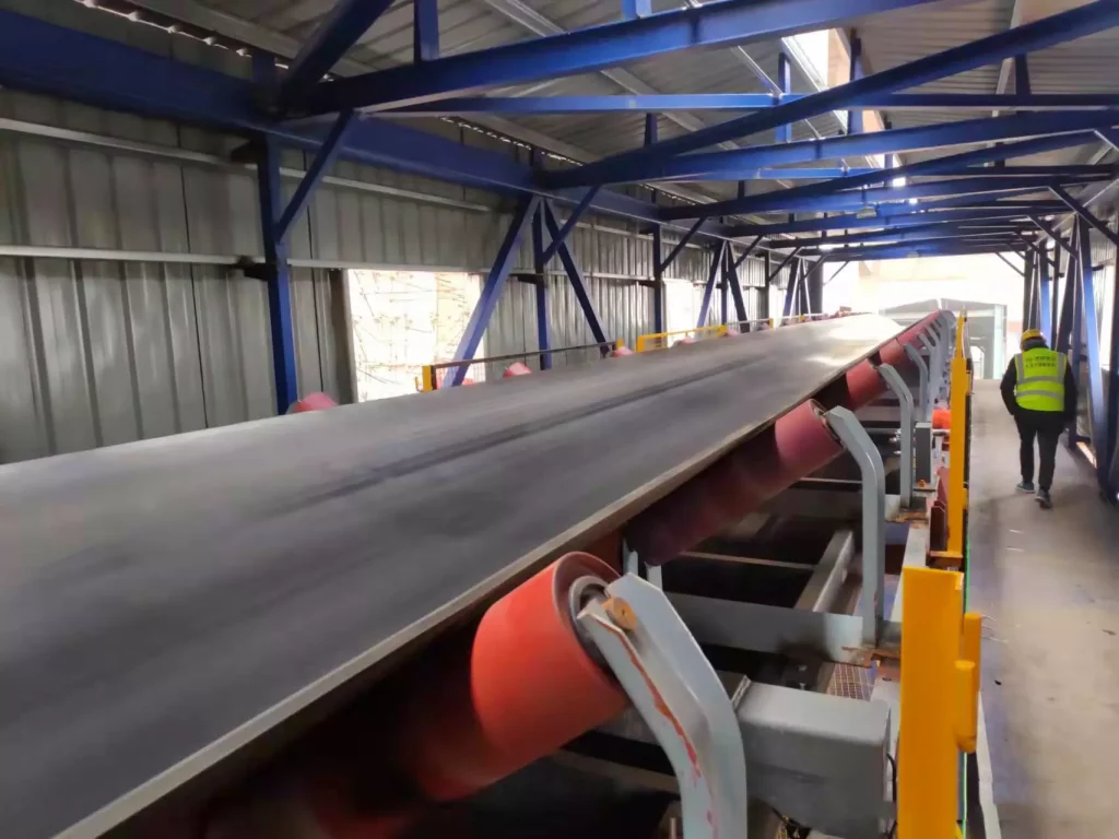 Practical Advice on Choosing Rollers or Belts for Industrial Weighing Line 36 A large industrial belt conveyor with red rollers and a black belt in a steel factory, used for continuous material transport, with a worker inspecting it.