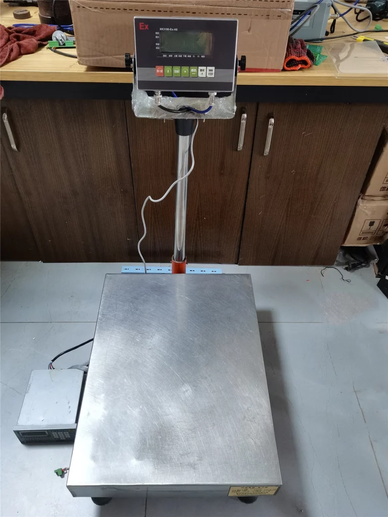 The Ideal Explosion-Proof Weighing Solutions for Harsh Environments 37 This is an explosion-proof bench scale. It has a stainless steel weighing platform and an indicator marked with "Ex" (model XK3150-Ex-A8) for weight display. Suitable for harsh environments like chemical plants and oil fields, it is situated in a workshop or laboratory area, with tools, cardboard boxes, and other items around.